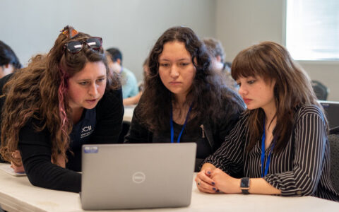 Three young women collaborate on a laptop, learning about cybersecurity careers at OCII and BSides Oklahoma event.