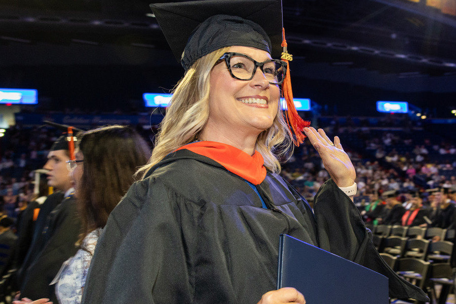 Woman in graduation cap and gown waves, holding diploma at Commencement 2026.