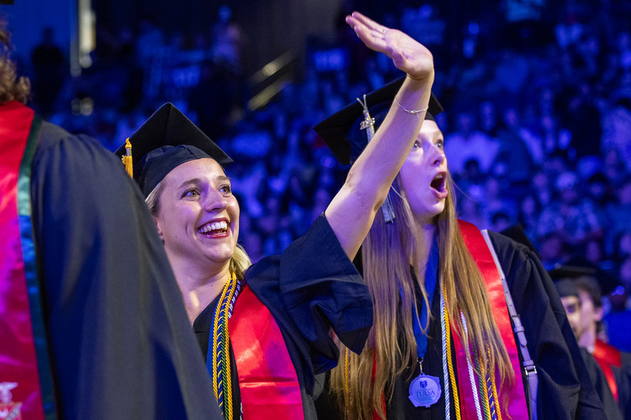 Graduate commencement 2026: Two smiling female graduates in caps and gowns wave to the crowd.