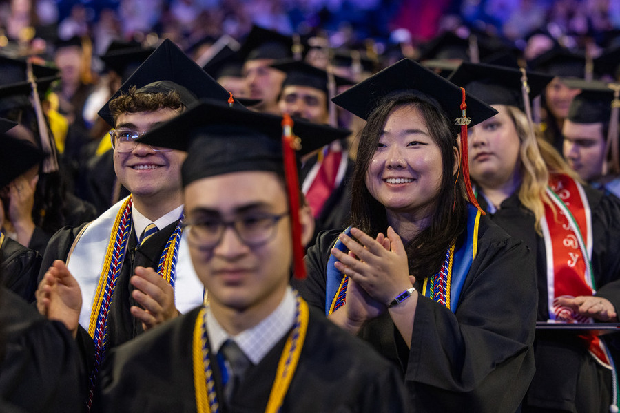 Smiling graduate in cap and gown applauds at 2026 commencement ceremony.