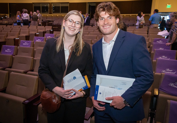 Two graduates in suits hold diplomas at a commencement ceremony