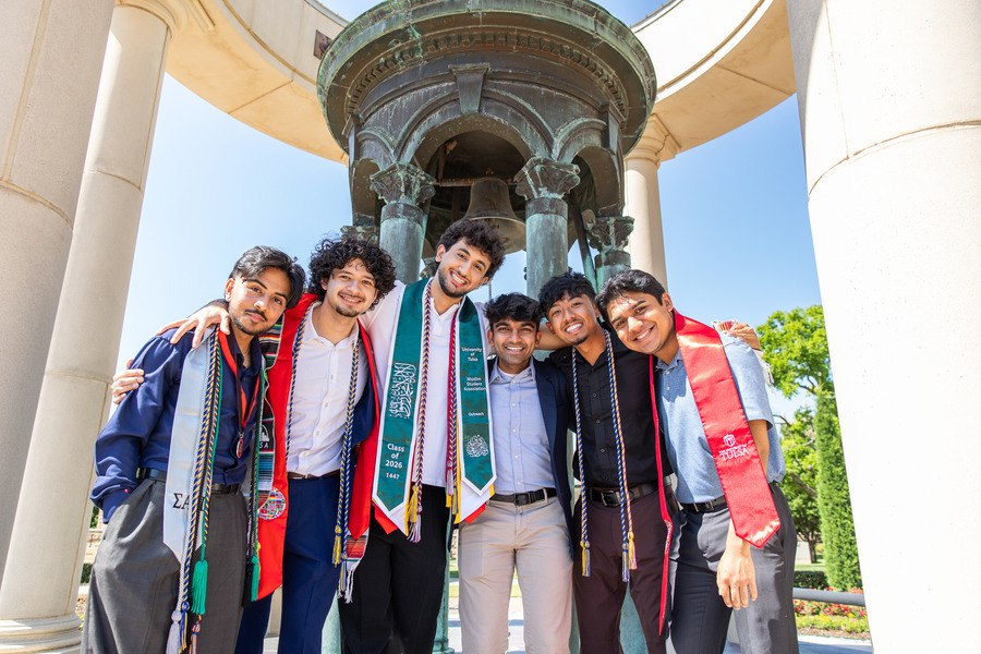 Six young men in graduation regalia, smiling in front of the Kendall Bell